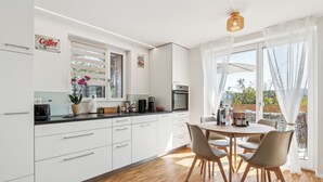 Une cuisine moderne avec des meubles blancs, un coin repas doté d’une table ronde et de chaises, et une vue sur l’extérieur grâce à de grandes fenêtres.
