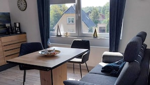 A modern living room with a sofa, a dining table, and a view of houses through a large window.