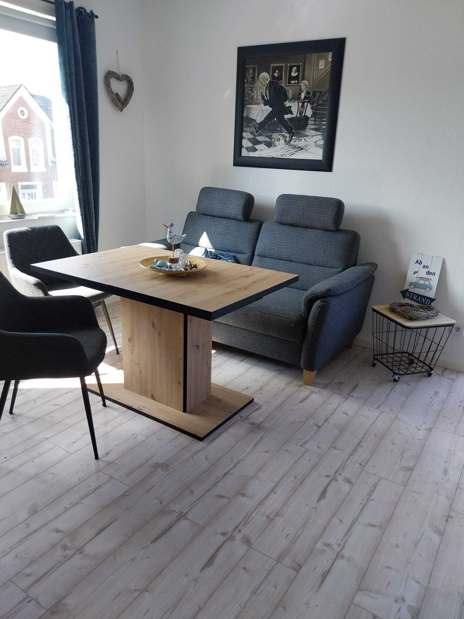 Ein modernes Wohnzimmer mit Esstisch aus Holz, grauem Sofa und einem gerahmten Bild an der Wand.