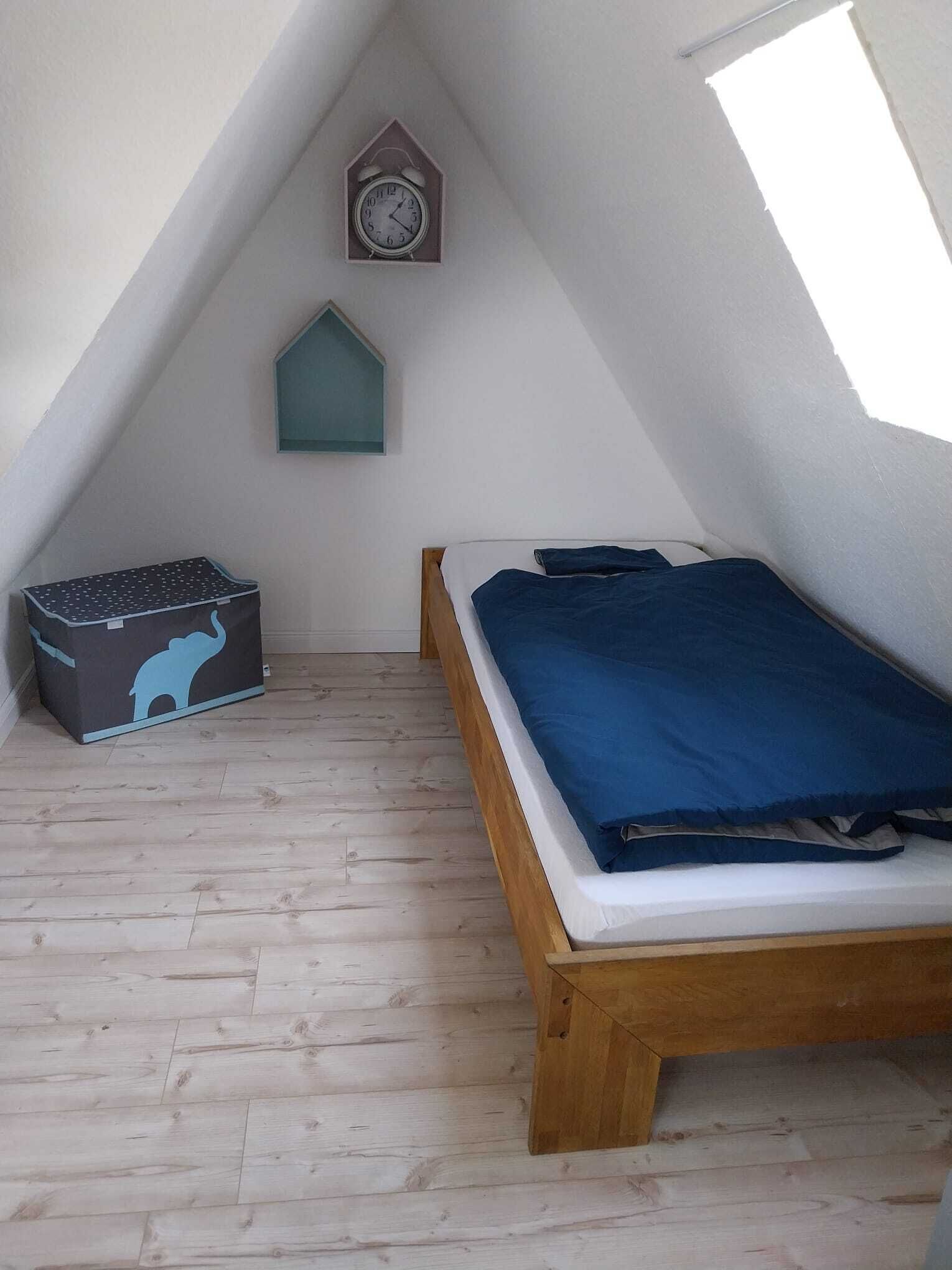 A small attic bedroom with a sloped ceiling, a wooden bed, a storage box, a clock, and a window.