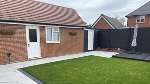 A residential backyard with a well-maintained lawn, a patio area with white marble tiles, a brick house with a white door, and a black fence.