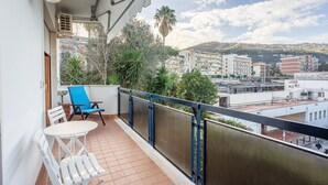 A balcony with a white table and blue chairs, overlooking a cityscape with buildings and a palm tree.