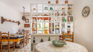 A dining area with a round table covered by a patterned tablecloth, surrounded by wooden chairs. A white shelving unit with various kitchen items and a window above the table is visible.