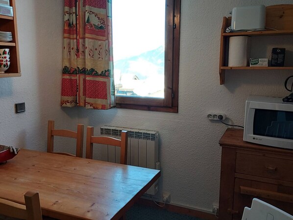 A small kitchen with a wooden table, a radiator, a microwave, and a window with a curtain.
