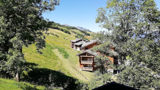 A wooden chalet with a balcony, surrounded by lush greenery and a clear sky.