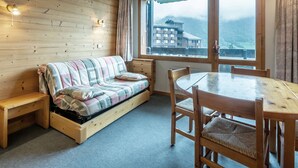 A cozy room with a wooden wall, a sofa with a striped blanket, a wooden table with chairs, and a large window with a view of buildings and mountains. - Cozy 2-room apartment in Valmorel, 50m from slopes, pets allowed (Valmorel)