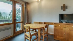 A cozy room with a wooden dining table, chairs, a television, and a large window offering a view of trees and mountains. - Cozy 2-room apartment in Valmorel, 50m from slopes, pets allowed (Valmorel)