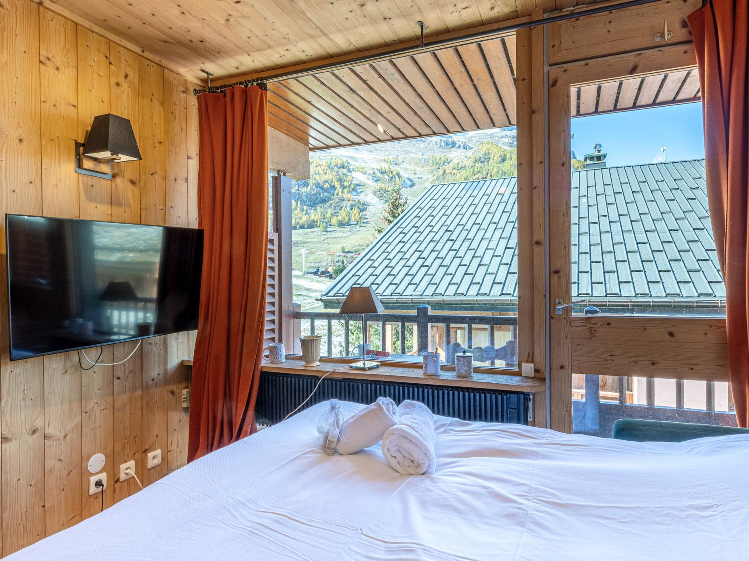 Ein Zimmer mit einem Bett, einem an der Wand befestigten Fernseher, einem Fenster mit Vorhängen und Blick auf ein Gebäude mit Ziegeldach.