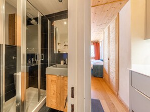 A modern bathroom with a glass shower enclosure, a wooden vanity, and a visible bed in the adjacent room.