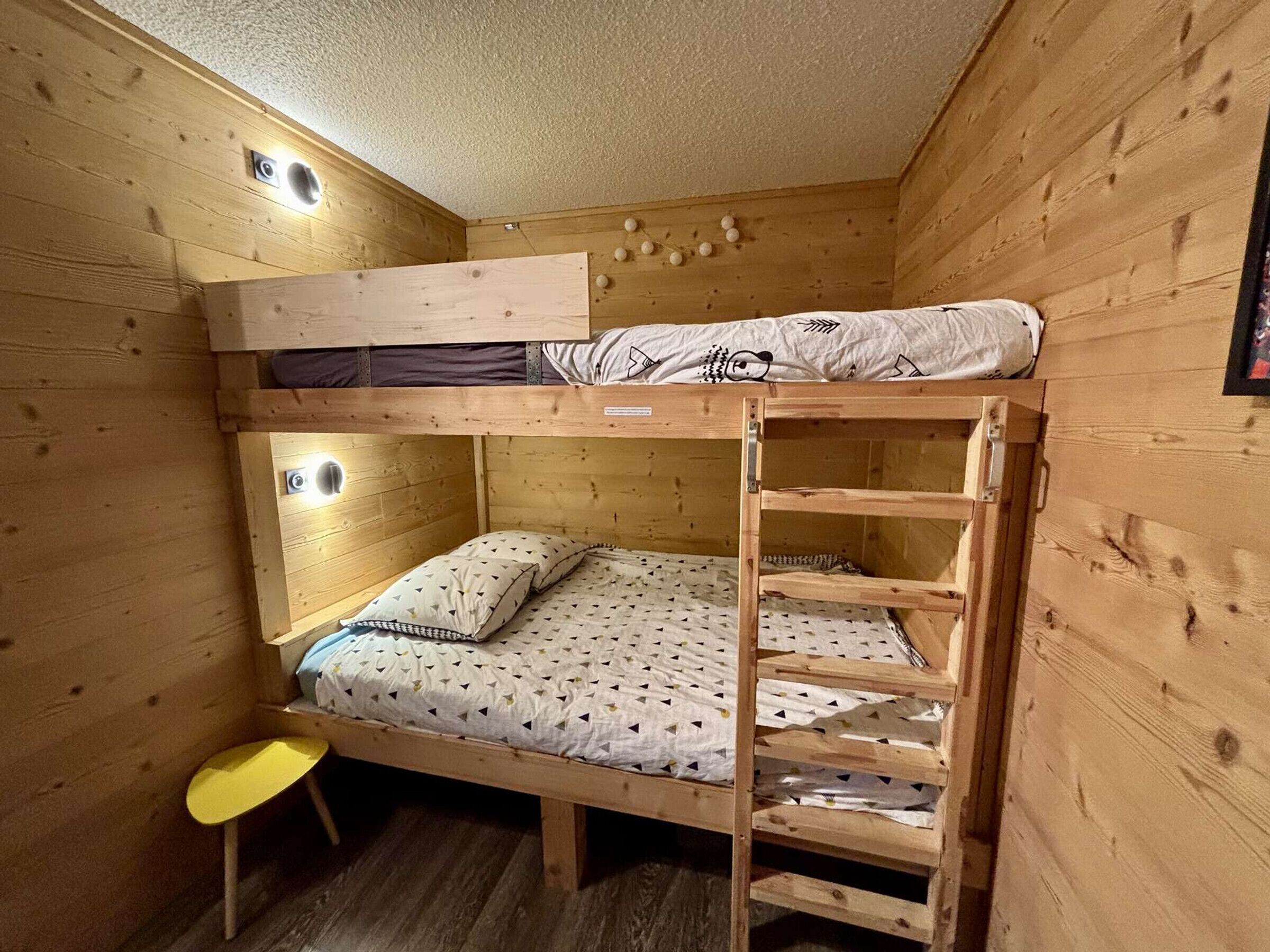 Ett rum med en våningssäng, väggar klädda med träpanel och en liten gul pall. En vägglampa sitter monterad på väggen.