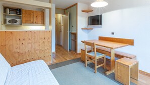 Une pièce de taille modeste comprenant un lit, un bureau en bois, une chaise, un petit tabouret, un téléviseur fixé au mur et un coin cuisine équipé d’un four à micro-ondes.