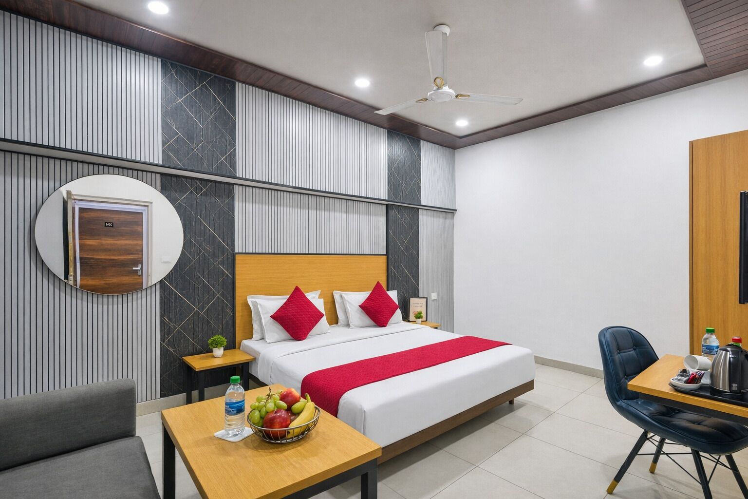 A modern hotel room with a bed, bedside table, chair, and a small table with a fruit bowl and water bottle.