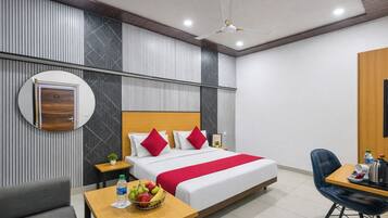 A modern hotel room with a bed, bedside table, chair, and a small table with a fruit bowl and water bottle.