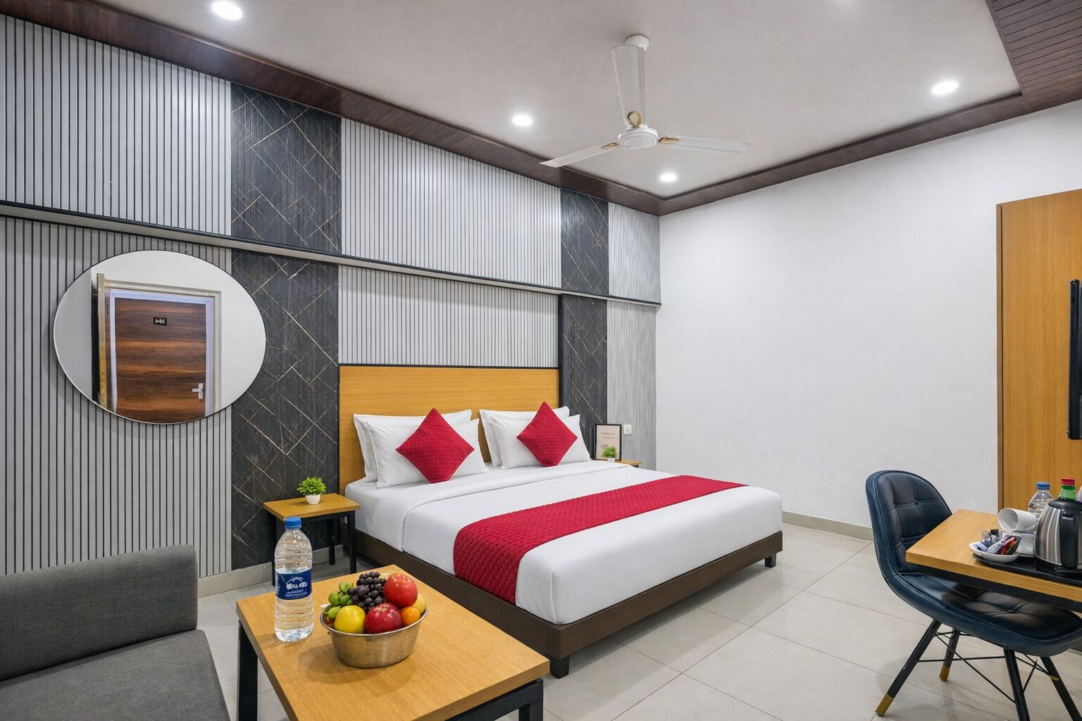 A modern hotel room with a bed, a round mirror, a bedside table with a water bottle, a fruit bowl, and a desk with a chair.