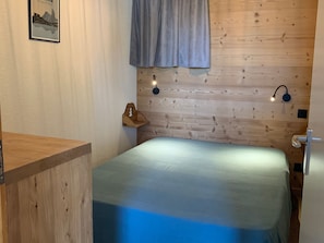 Une chambre à coucher avec une tête de lit en bois, un lit recouvert d’une couvre-lit bleue, deux lampes de lecture fixées au mur, une table de chevet en bois et un tableau encadré accroché au mur.