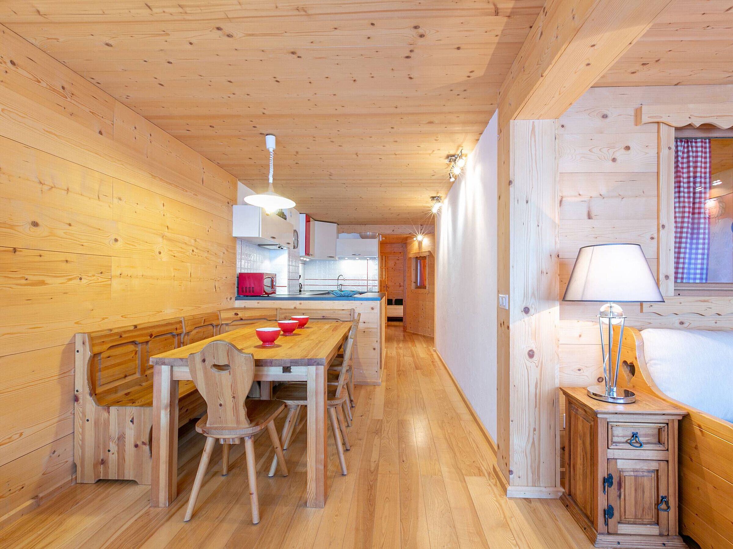 A wooden interior with a dining area, kitchen, and a small bedside table with a lamp.
