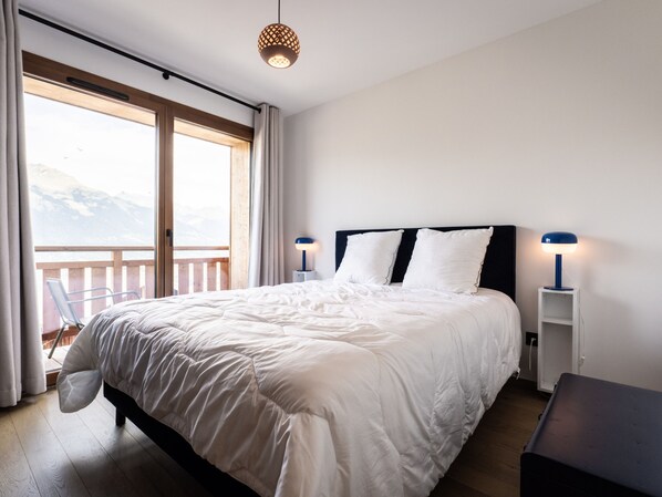 A bedroom with a large bed, two bedside lamps, a view of mountains through a window, and a balcony.