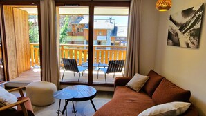 A cozy living room with a brown sofa, a small round table, and two chairs. A large sliding glass door leads to an outdoor deck with a wooden railing and two chairs. - Beautiful apartment in Plagne Montalbert, near slopes with wifi and parking (La Plagne Montalbert)