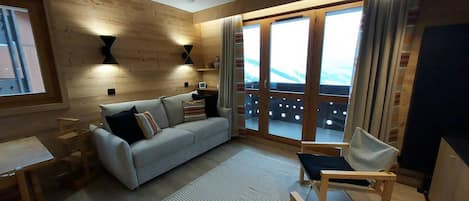 Ein modernes Wohnzimmer mit einer Couch, einem Couchtisch und einem Sessel. Ein Balkon bietet Blick auf die Berge.