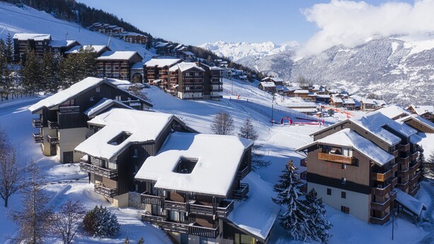 Een bergresort bedekt met sneeuw, met houten gebouwen, skipistes en een helderblauwe lucht.