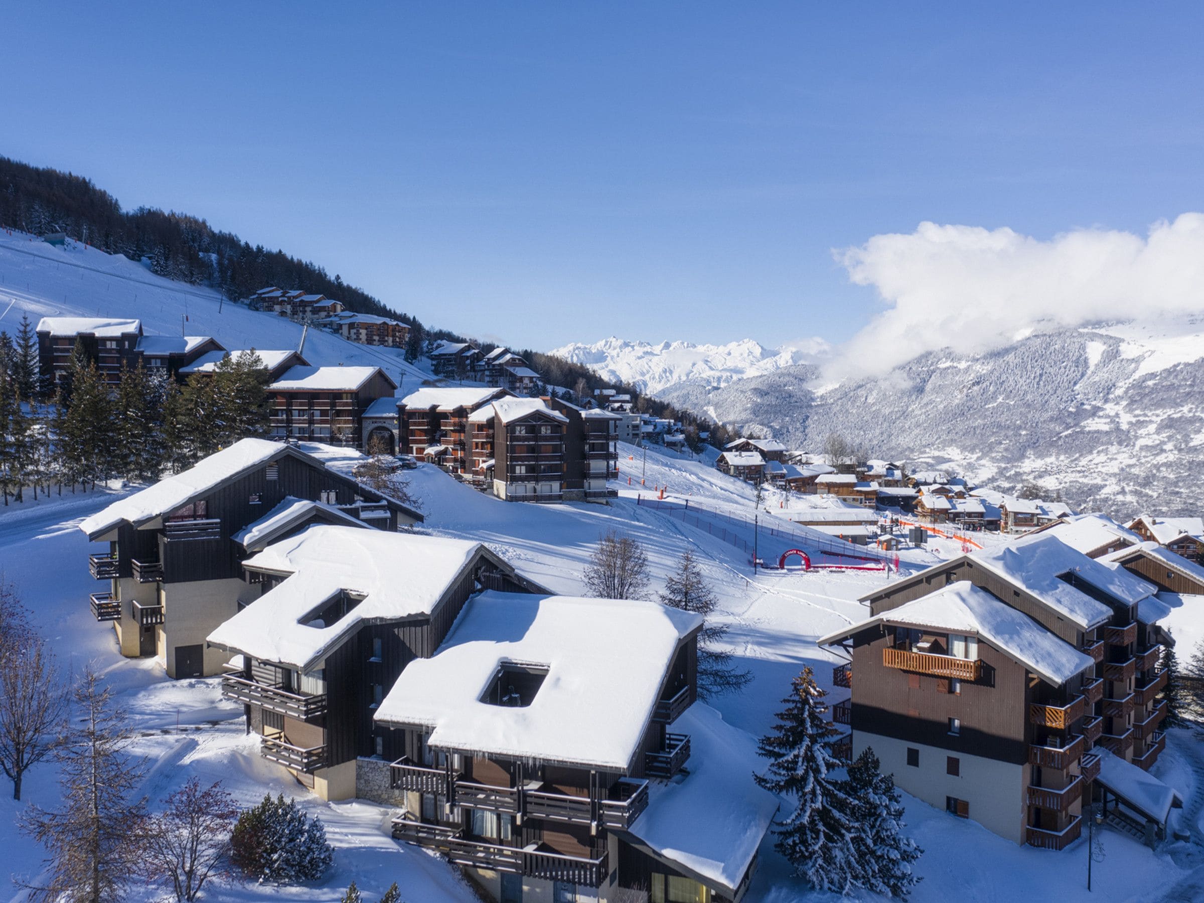 A snow-covered mountain resort with wooden buildings, ski slopes, and a clear blue sky.