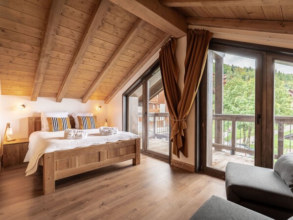 Un dormitorio con estructura de madera, una cama grande, una mesita de noche, una lámpara y vistas a un balcón y follaje exterior.