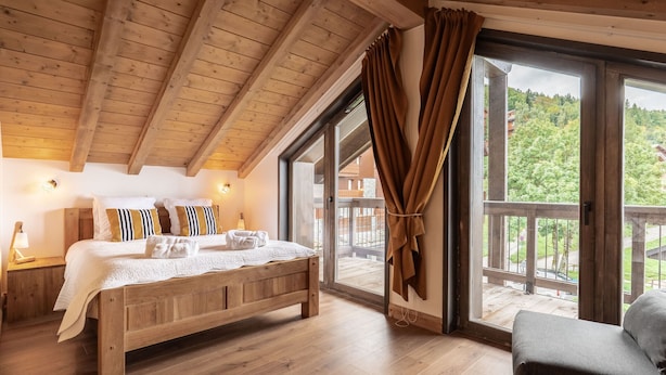 Een slaapkamer met houten frame, met een groot bed, een nachtkastje, een lamp en uitzicht op een balkon en groen buiten.