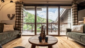 Een gezellige woonkamer met een houten vloer, twee gestreepte banken, een ronde houten tafel met twee kopjes erop en een grote schuifdeur die leidt naar een balkon met uitzicht op een bebost gebied.