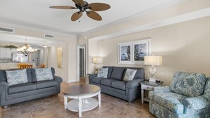 A spacious living room with two grey sofas, a round coffee table, and a patterned armchair. A ceiling fan and artwork are visible on the walls. - Sandy Toes Paradise Retreat: 2BR Oceanfront Condo (North Myrtle Beach)