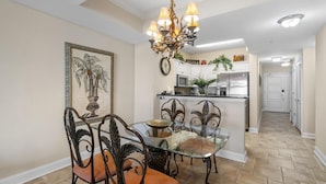 Un comedor con mesa de vidrio, sillas ornamentadas, una araña y una cocina al fondo.