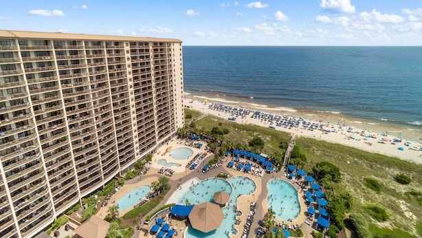 Un hotel de gran altura con varios balcones, un área de piscina frente a la playa y vistas al océano.