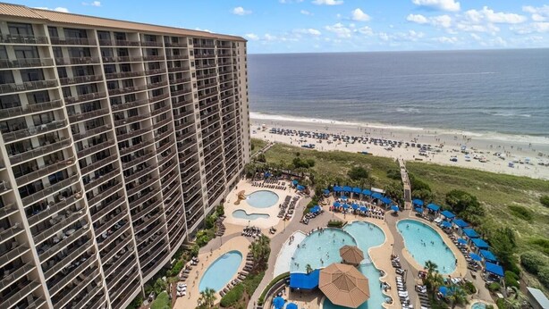 Un hotel de gran altura con múltiples balcones con vistas a una playa y un área de piscina con varias piscinas y tumbonas.