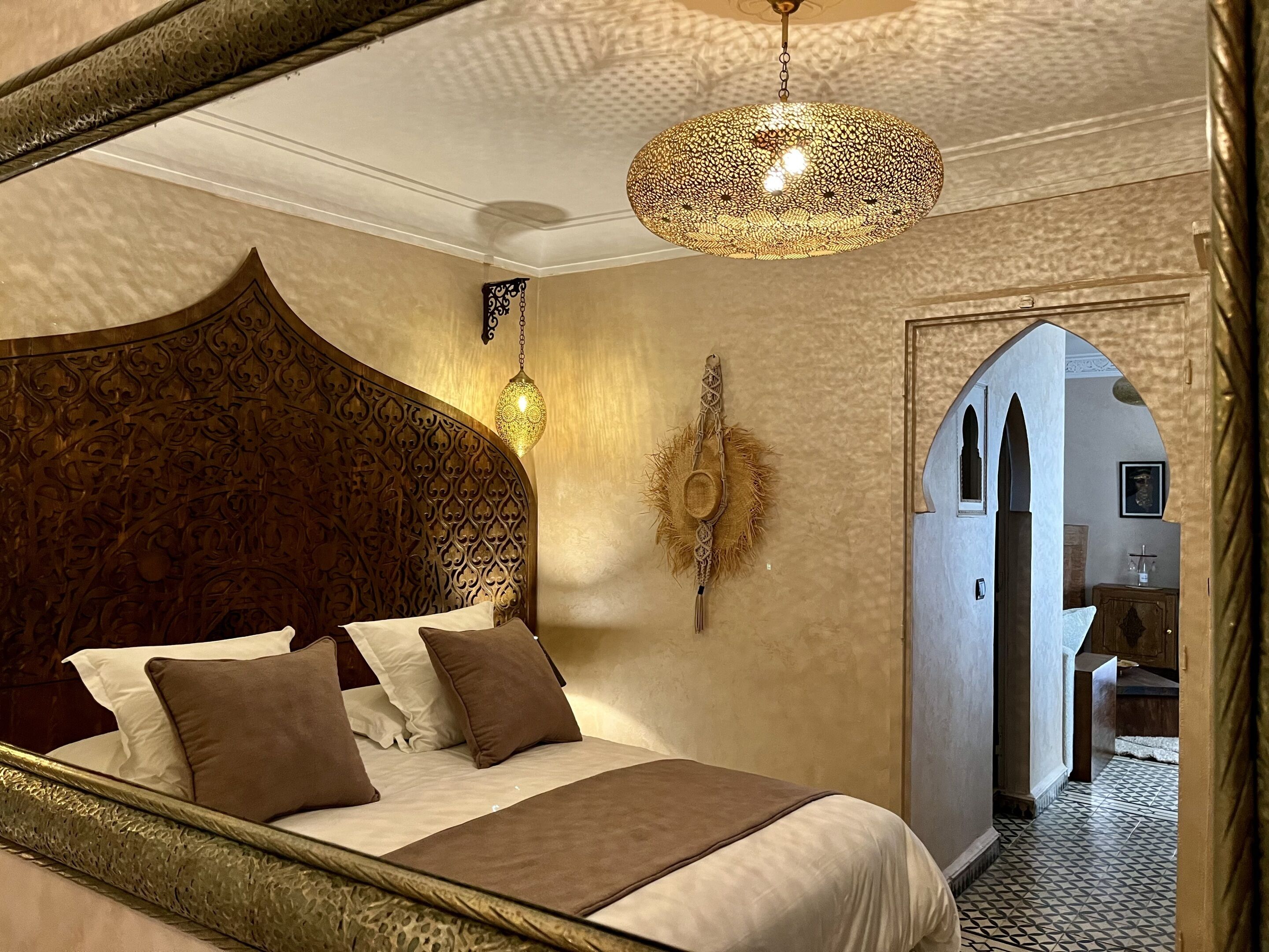 A bedroom with a large bed, ornate headboard, a hanging lantern, and a view of another room through an arched doorway.