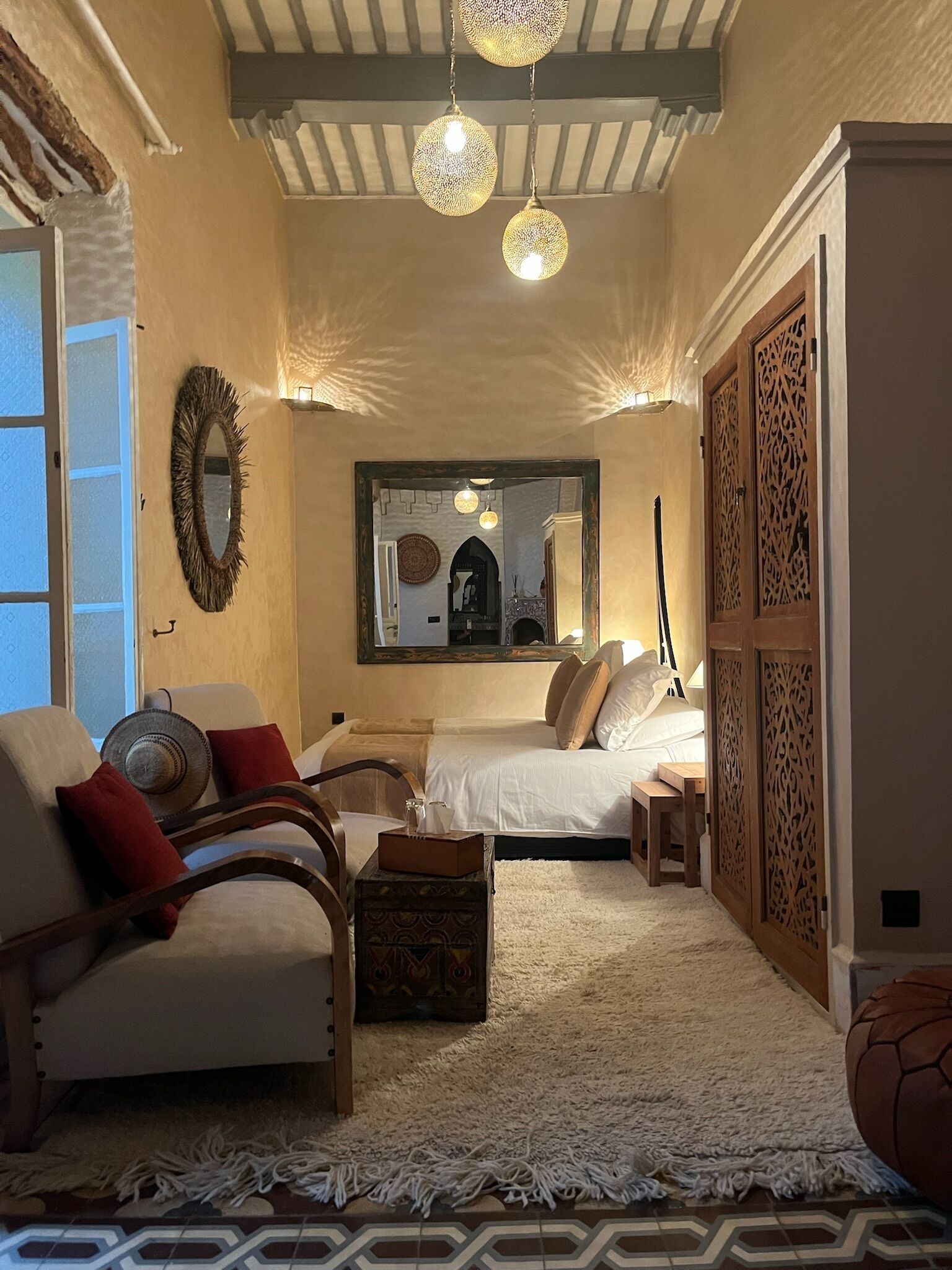 A cozy bedroom with a bed, a chair, a small table, and a decorative mirror.
