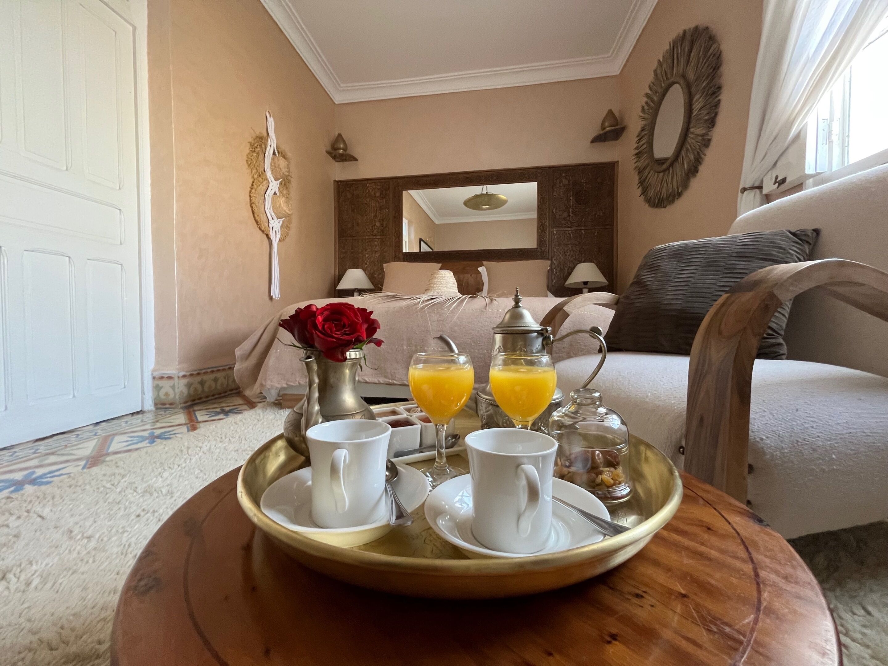 Ein Frühstücksbrett mit Tassen, einer Karaffe und Orangensaft auf einem runden Tisch in einem Zimmer mit Bett, Spiegel und Fenster.
