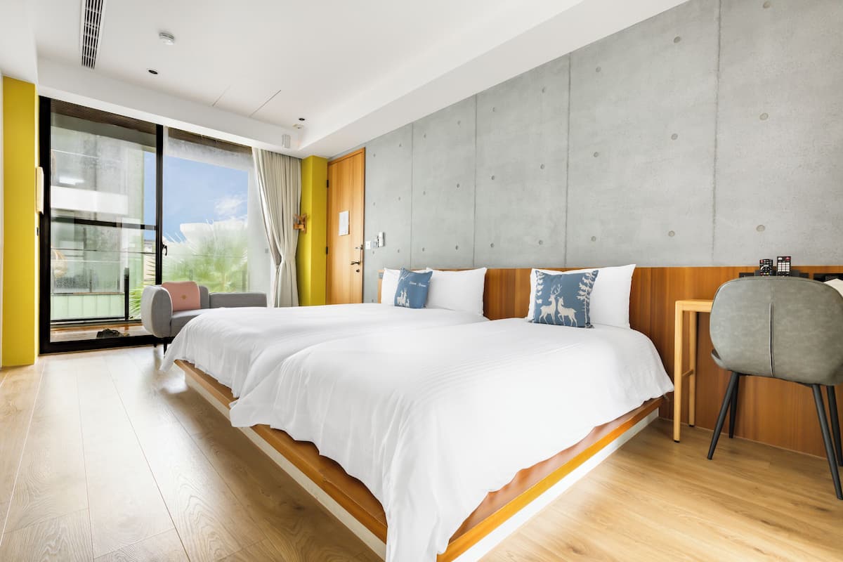 A modern hotel room with a large bed, wooden flooring, a concrete accent wall, and a view through a glass door.