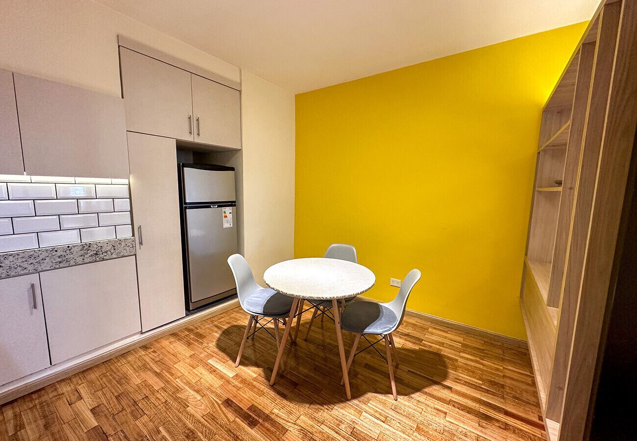 Une cuisine de taille réduite comprenant un réfrigérateur, une table à manger avec des chaises et un mur d’accent jaune.