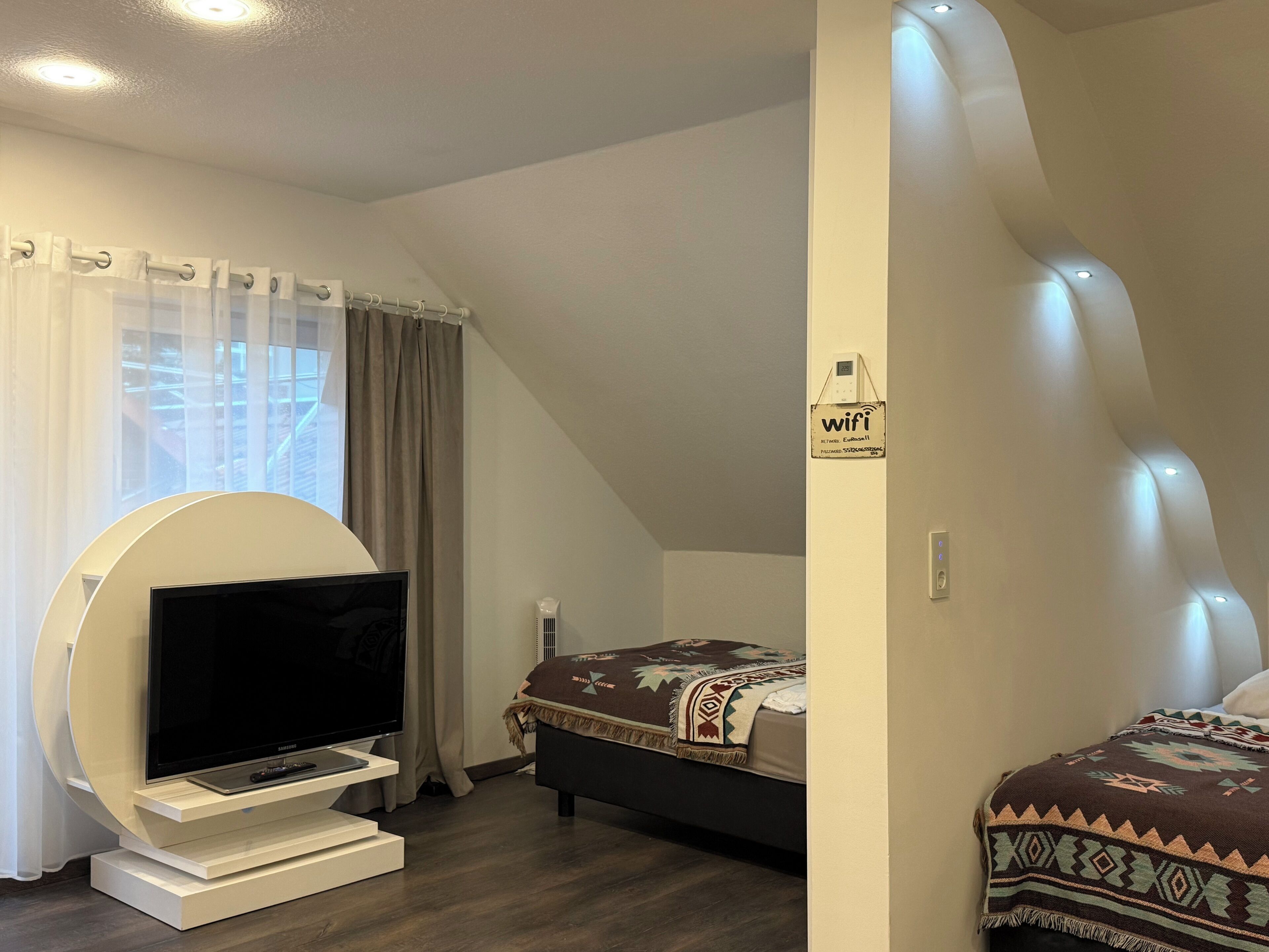 Een moderne slaapkamer met een televisie op een witte standaard, een bed met een gebloemd deken en een 'wifi'-bordje aan de muur.