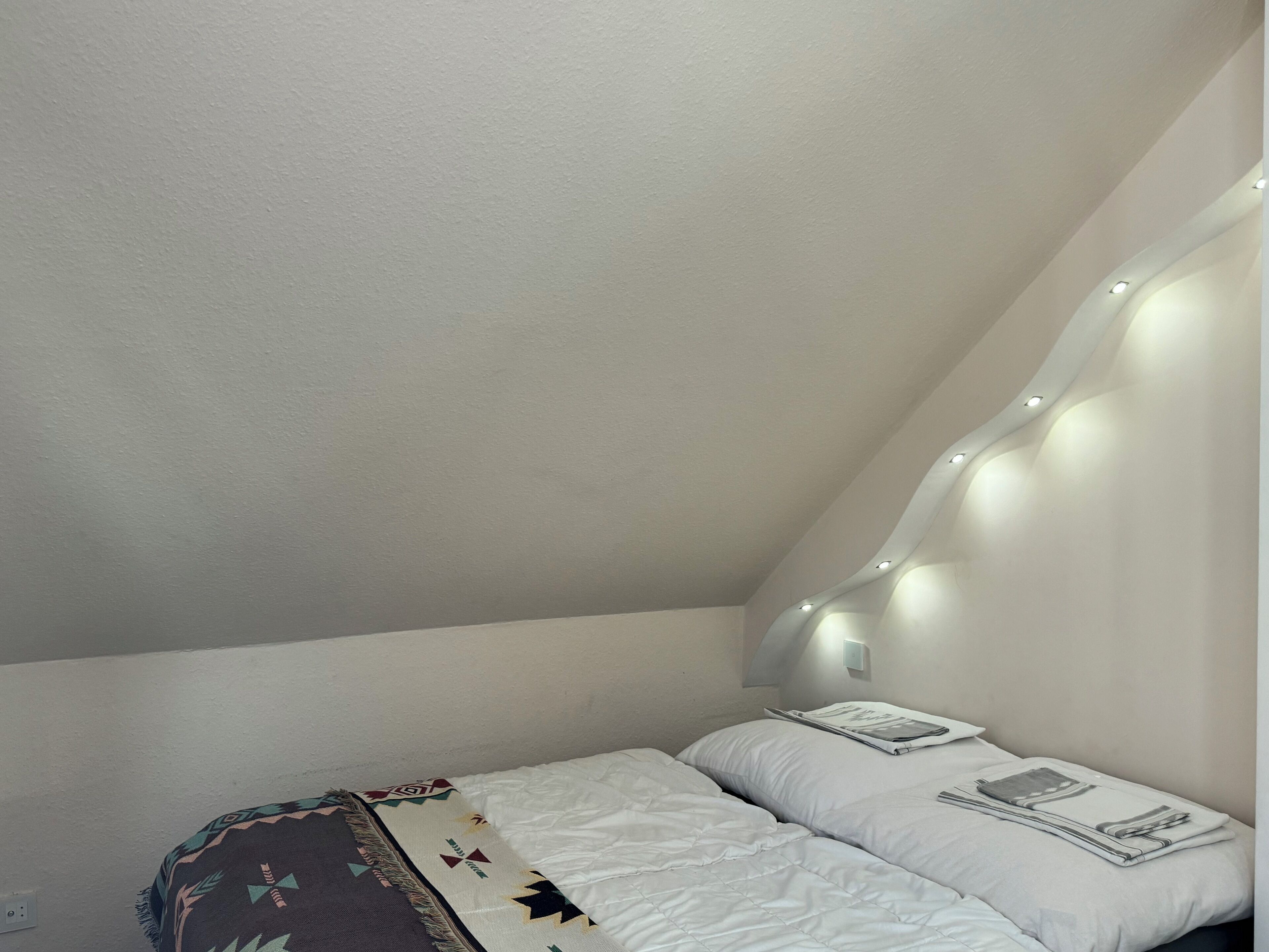 A bedroom with a sloped ceiling, a bed with white bedding, a patterned throw blanket, and wall-mounted lights.
