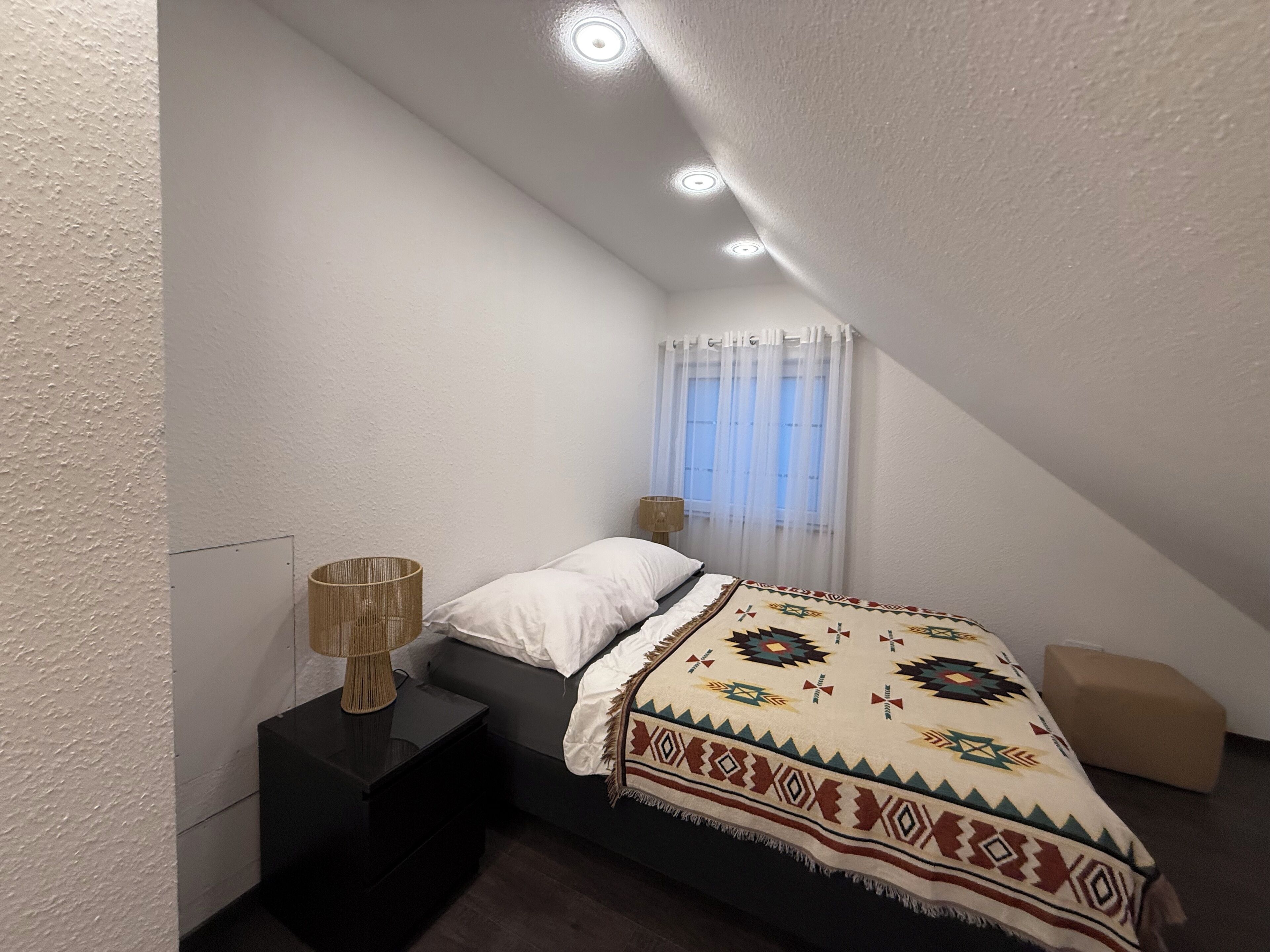 A bedroom with a sloped ceiling, a bed with a patterned blanket, a bedside lamp, and a window with sheer curtains.