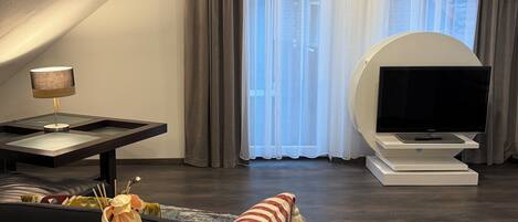 Una sala de estar moderna con un televisor de pantalla plana sobre un soporte blanco, una mesa de centro de vidrio, un sofá de cuero negro y un jarrón decorativo con flores.