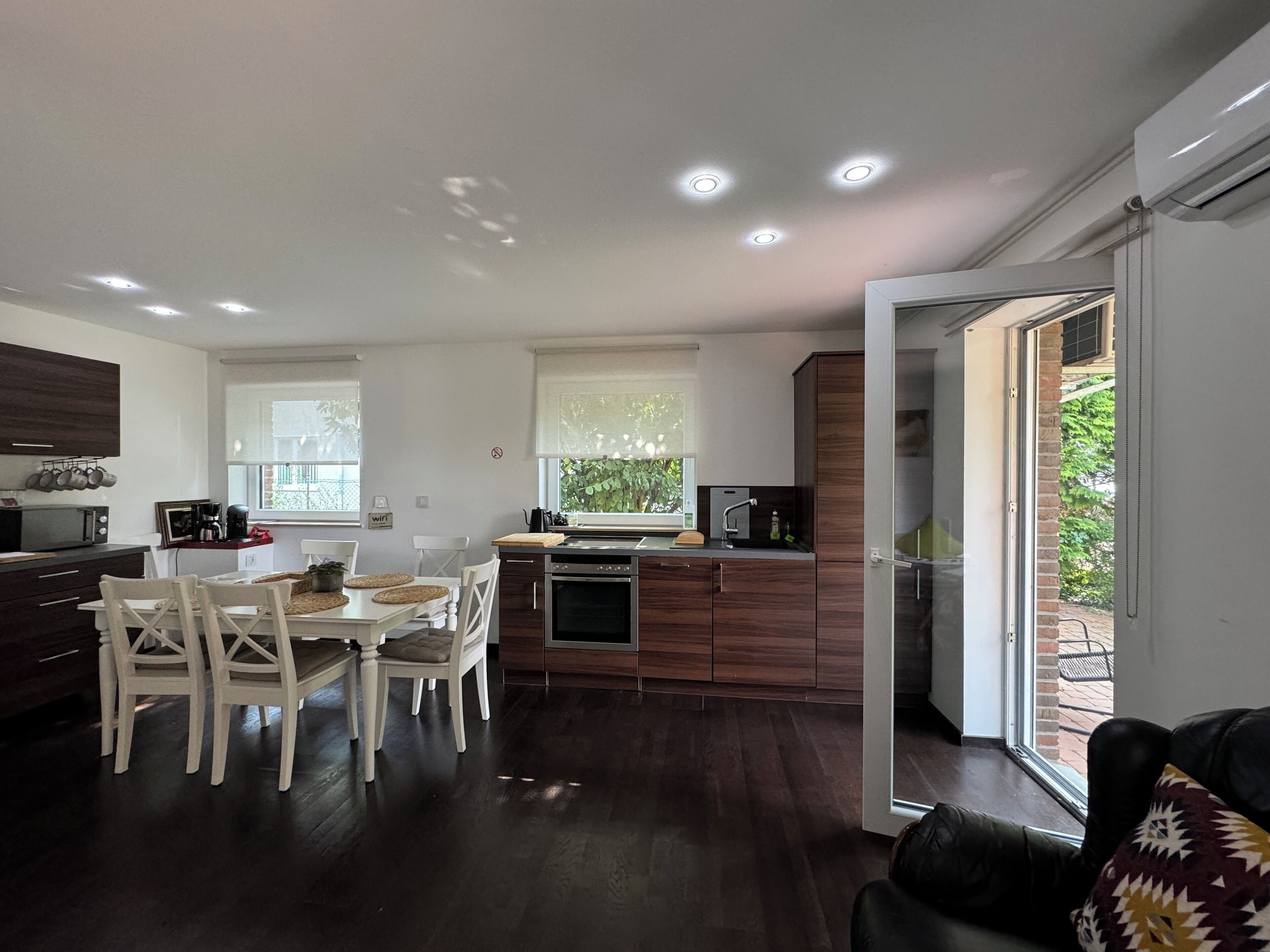 A modern kitchen with dark wood cabinets, a dining area with white chairs, and a large window providing natural light.