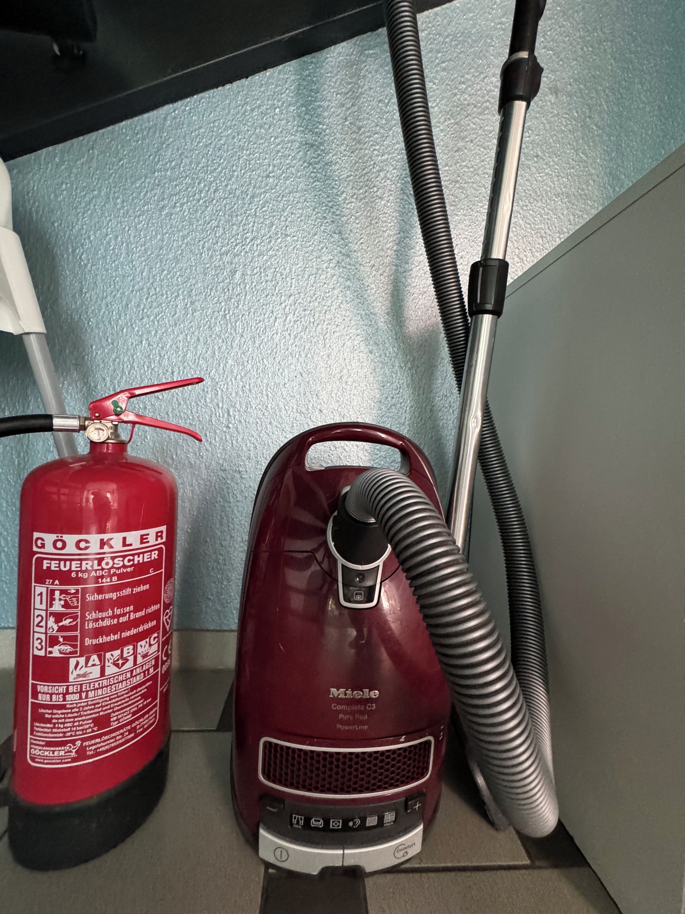 A red fire extinguisher and a vacuum cleaner with hoses, placed against a wall.