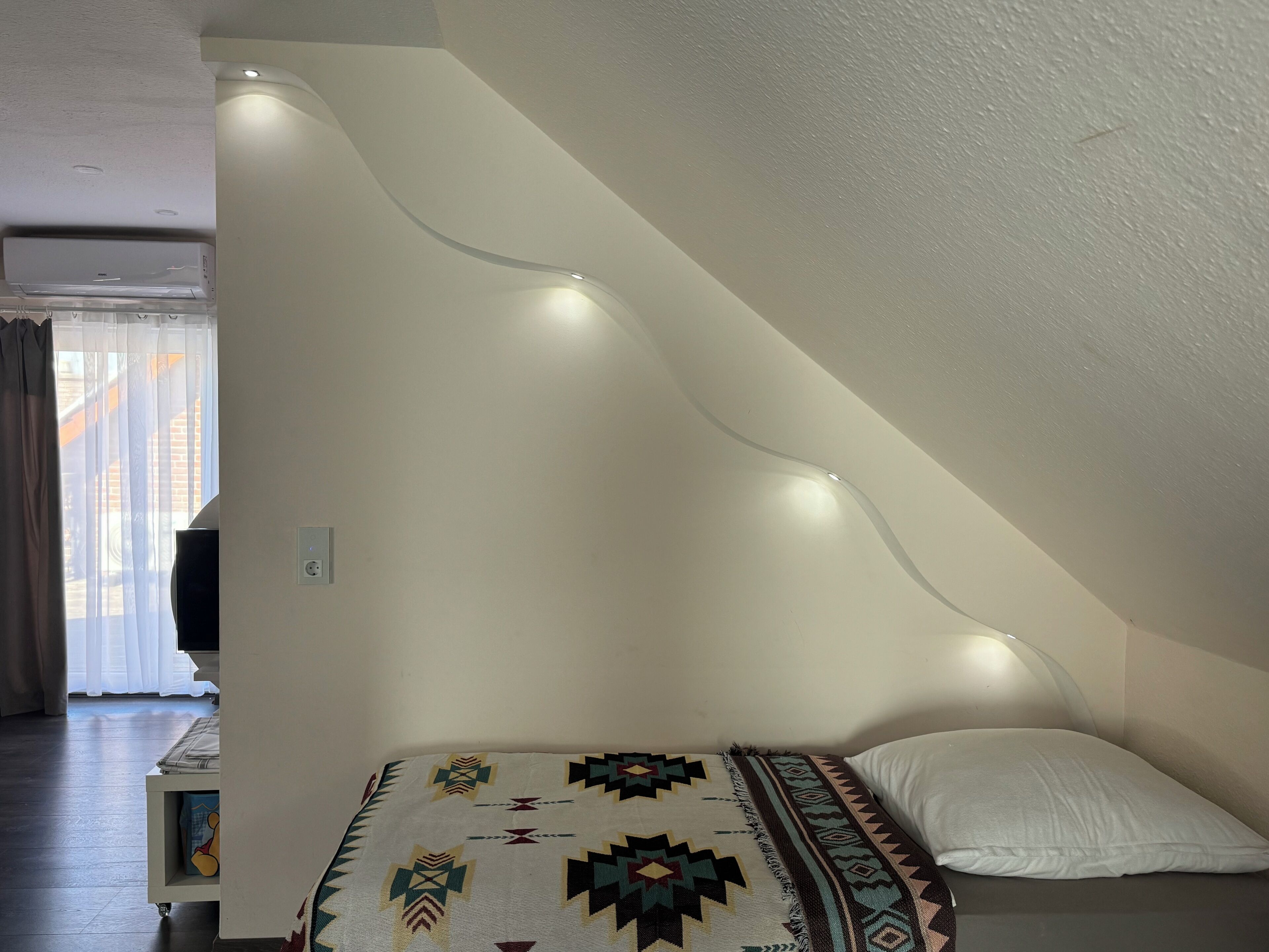 Een slaapkamer met een schuine plafond, een bed met een geborduurd plaid, een wit kussen, en een wandlamp.