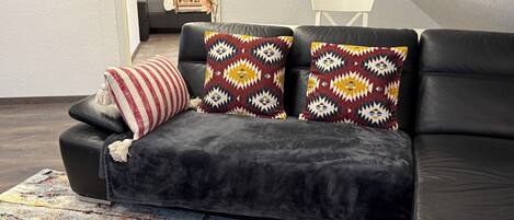 Un sofá de cuero negro con cojines estampados, una alfombra colorida y una silla blanca en una habitación moderna con una ventana y una puerta.