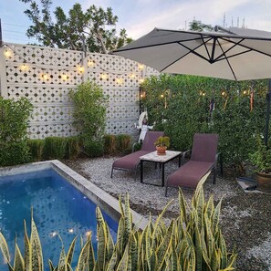 A poolside area with a small pool, two lounge chairs, a table with a potted plant, and a decorative wall with string lights.
