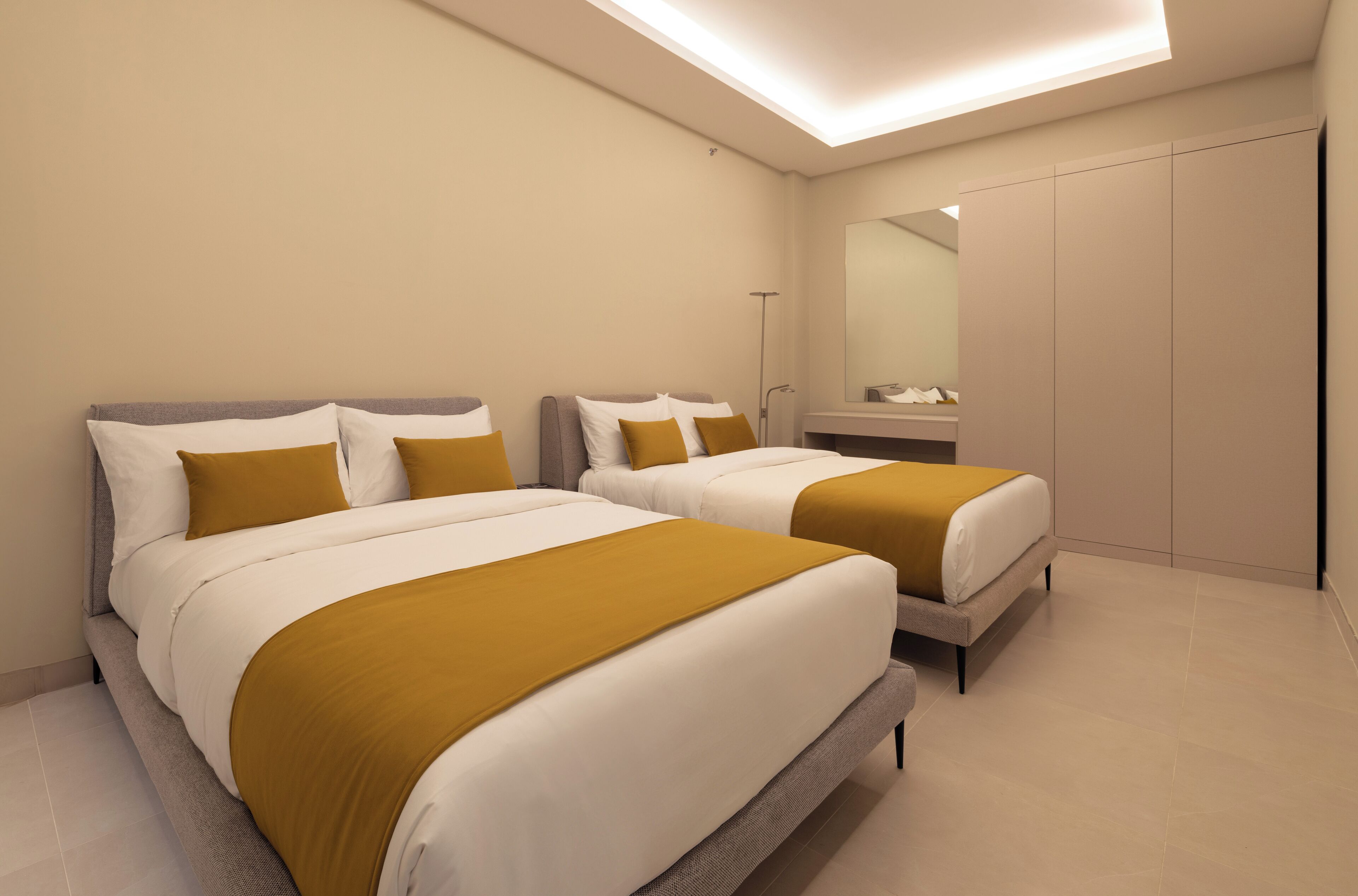 Ein modernes Hotelzimmer mit zwei Betten, jeweils mit weißer und senffarbener Bettwäsche, einem großen Spiegel und einem begehbaren Kleiderschrank.