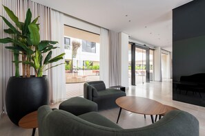 A modern interior with a large potted plant, grey upholstered chairs, a wooden table, and a view of the outdoors through large windows.