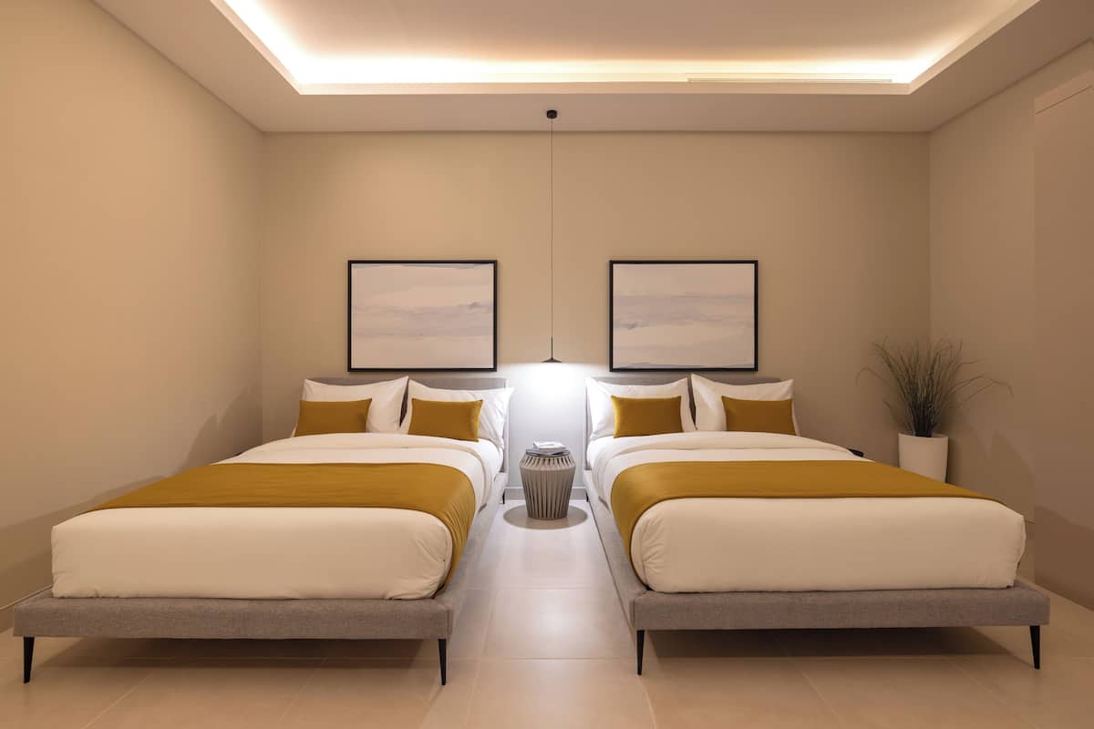 Moderni hotellihuone, jossa on kaksi sänkyä, keskeisellä paikalla oleva yöpöytä ja seinillä tauluja.