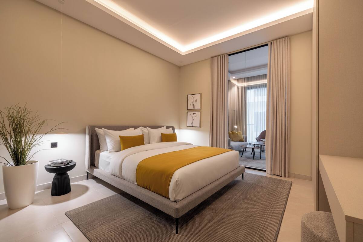 Moderni hotellihuone, jossa on suuri sänky, yöpöytä, ruukkukasvi ja näkymä viereiseen huoneeseen.